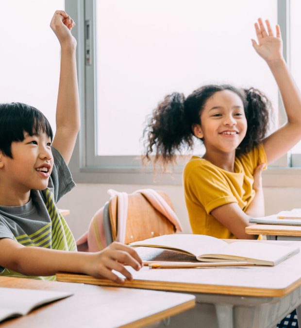 Elementary age Asian student boy raised hands up in class. Diverse group of pre-school pupils in elementary age in education building school. Volunteering and participating classroom concept.