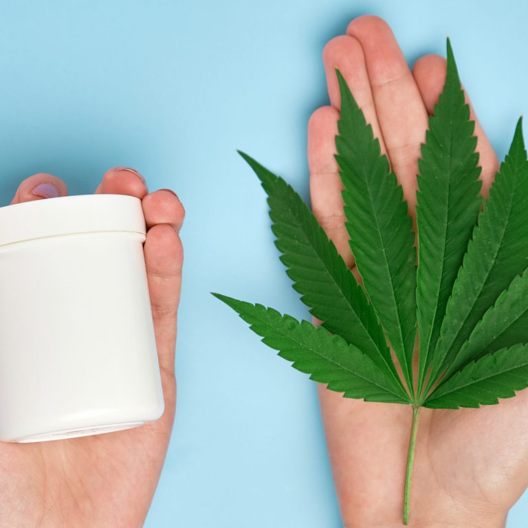 Close up woman hands holding a package with cream with copy space and marijuana leaf on the blue background. Skin care or dermatology treatment with cannabis hemp oil.